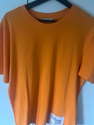 Orange t-shirt från Zara - Snygg orange t-shirt från Zara med rund hals och korta ärmar. Tillverkad i mjuk bomull och har en liten vit patch med text nere vid fållen. Perfekt för dig som gillar färgstarka basplagg.