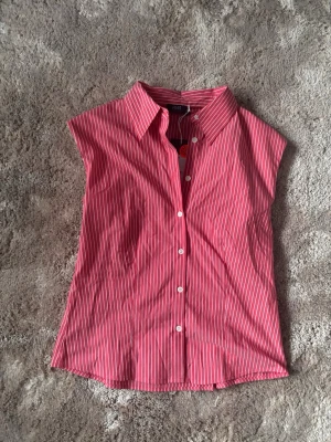 Rosa randig skjorta från Vost - Kortärmad skjorta från Vost i rosa med vita ränder. Klassisk krage och knäppning framtill med vita knappar. Skjortan har en avslappnad passform och är tillverkad i ett lätt bomullstyg, perfekt för varmare dagar.