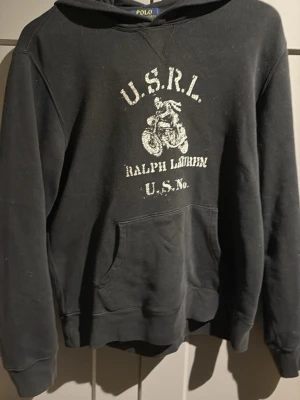 Svart hoodie från Ralph Lauren - Snygg svart hoodie från Polo Ralph Lauren med tryck på bröstet som visar en motorcykel och texten 'U.S.R.L. Ralph Lauren U.S. No.'. Klassisk känguruficka framtill och huva. Perfekt för en avslappnad streetwear-look.   Passar M