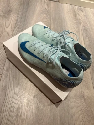 Nike Mercurial blå fotbollsskor - Snygga Nike Mercurial fotbollsskor i ljusblått med blå swoosh och detaljer. Skorna har låg profil, snörning och är tillverkade i ett lätt syntetmaterial för bästa känsla på planen. Perfekta för dig som vill ha speed och kontroll på konstgräs eller gräs. Priset går och diskutera!