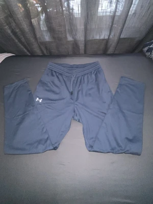 Mörkblå mjukisbyxor från Under Armour - Snygga mörkblå mjukisbyxor från Under Armour med dragsko i midjan och elastisk resår. Byxorna har en enkel design med logga på vänster lår och är tillverkade i ett mjukt och stretchigt material som känns skönt mot huden.