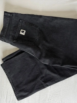 Svarta Carhartt jeans med rak passform - Svarta jeans från Carhartt med rak passform och klassisk femficksdesign. Jeansen har en tydlig Carhartt-logga på bakfickan och är tillverkade i kraftig denim. Perfekta för en avslappnad streetwear-look.