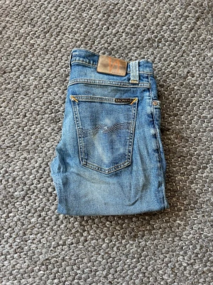 Nudie Jeans - Säljer ett par blå skinny jeans från Nudie Jeans med klassisk femficksdesign och snygga slitningar. Jeansen har en ljus tvätt och detaljerade sömmar på bakfickan. Perfekta för en avslappnad och den ljusa färgen gör plagget lämpligt för vår och sommar.