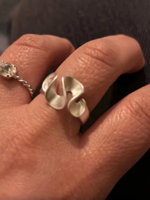 vågig ring design - Cool  ring i silver metall med vågig, abstrakt form. Ringen har en bred och öppen design som verkligen sticker ut och ger en modern känsla. Perfekt accessoar för dig som gillar trendiga och iögonfallande smycken.