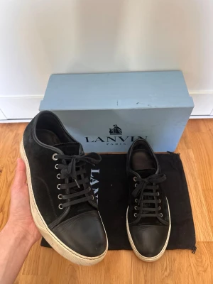 Svarta Lanvin sneakers i mocka och läder - Snygga svarta sneakers från Lanvin med ovandel i mocka och tåparti i slätt läder. Skorna har vita gummisulor, svarta skosnören och metallfärgade öljetter. Klassisk låg modell som passar till det mesta. Kommer med originalkartong och dustbag.