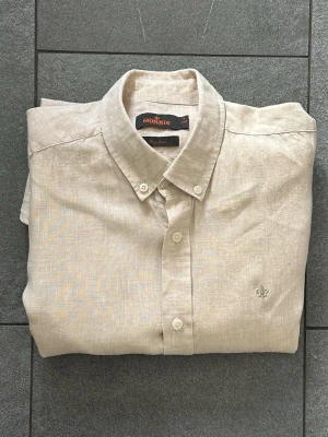 Beige linneskjorta från Morris - Stilren beige skjorta från Morris i linne med klassisk krage och knäppning framtill. Skjortan har lång ärm och en liten broderad logga på bröstet. Perfekt för en avslappnad men snygg look.
