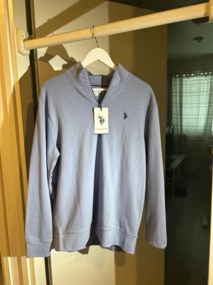 Ljuslila hoodie från U.S. Polo Assn. - Snygg ljuslila hoodie från U.S. Polo Assn. med dragkedja vid halsen och broderad logga på bröstet. Tillverkad i mjukt material och har långa ärmar samt ribbade muddar. Perfekt för en avslappnad stil.