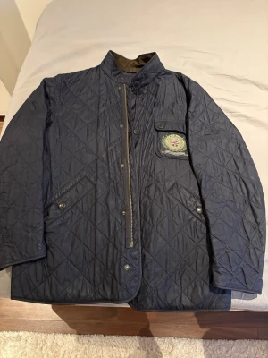 Marinblå quiltad jacka från Barbour - Snygg marinblå quiltad jacka från Barbour med klassisk krage i manchester och patch med brittisk flagga på bröstet. Jackan har dragkedja och knappar framtill, samt flera fickor. Perfekt för dig som gillar tidlös och stilren design.
