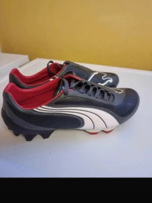 Svarta Puma fotbollsskor med röd insida - Säljer ett par snygga fotbollsskor från Puma i svart med vit logga och detaljer samt röd insida. Skorna har klassisk snörning och fasta dobbar för spel på gräs. Tillverkade i slitstarkt syntetmaterial som ger bra bollkontroll och passform.