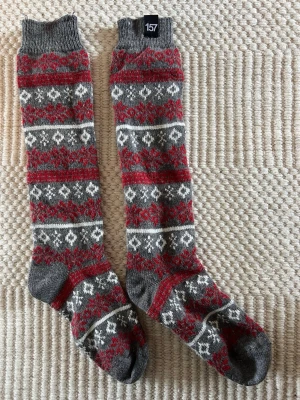 Grå och röda mönstrade långa sockor - Säljer ett par långa varma sockor i grått, rött och vitt med klassiskt vintermönster från lager157. Sockorna har ribbad kant upptill och är perfekta för kalla dagar. Storlek 36-40 enligt etiketten. Materialet känns mjukt och värmande, troligen ullblandning.