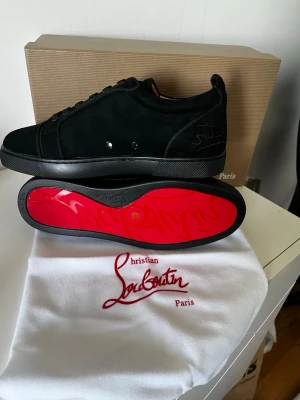 Svarta sneakers med röd sula - Snygga svarta sneakers med klassisk röd sula och diskret logga på hälen. Skorna är låga och har en stilren design i mocka och läder, perfekta för dig som vill sticka ut med exklusiva detaljer. Levereras med originalkartong och dustbag. Få ett valfritt bälte om du betalar 100kr extra!