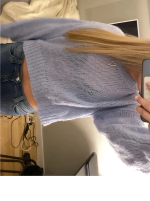 Ljusblå stickad croppad tröja - Säljer en ljusblå stickad tröja med croppad passform och långa ärmar. Tröjan har en ribbad kant vid ärmslut och nederkant samt ett litet hjärtdetalj vid halsen. Perfekt för dig som gillar en avslappnad och trendig stil.
