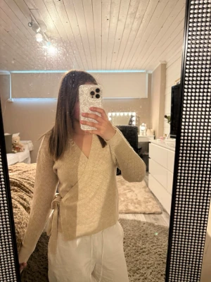 Beige omlottstickad tröja - Säljer en beige stickad omlottopp med knytband i sidan och diskret mönster. Tröjan har v-ringning och långa ärmar. Säljs pga: använder inte 