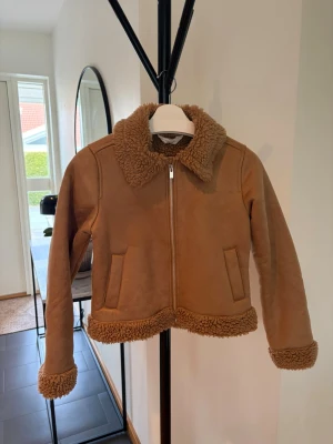 Brun teddyjacka med dragkedja - Snygg brun teddyjacka med mjukt foder och detaljer i teddy på krage, ärmslut och nederkant. Jackan har två fickor framtill och stängs med dragkedja. Perfekt för kyliga dagar och ger en trendig look.