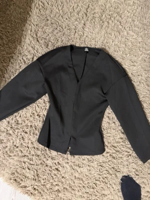 Svart figursydd kavaj - Säljer en svart figursydd kavaj från Shein med djup V-ringning och långa ärmar. Kavajen har en minimalistisk design utan knappar eller fickor och är tillverkad i ett slätt tyg som ger en stilren look. Säljs pga: gillar inte att man kan se huden 
