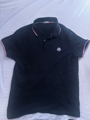 Svart Moncler pikétröja  - Snygg svart pikétröja från Moncler med klassisk krage och knappar framtill. Tröjan har Moncler-logga på bröstet och vita samt röda detaljer på krage och ärmslut. Perfekt för en stilren och sportig look.                                                               OBS: Pris kan diskuteras, om du har några frågor så är det bara att skriva.