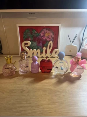 Söt parfymkollektion med unika flaskor - Samling med sex parfymer i olika färgglada och lekfulla flaskor, bland annat en rosa flaska med guldkrona, ett rött äpple och en flaska med rosa rosett. Flera flaskor har pastellfärger och fantasifulla former. Perfekt för dig som gillar att samla på snygga parfymflaskor.