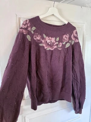 Lila stickad tröja med blommor Newbie - Lila stickad tröja från Newbie med strukturstickade prickar och blommigt mönster i rosa och grönt över bröstet och axlarna. Tröjan har rund halsringning, långa ärmar och ribbade muddar. Perfekt för dig som gillar romantisk och mysig stil. Passar snarare S/M.