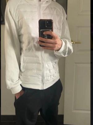 Vit quiltad pufferjacka med huva - Snygg vit pufferjacka med quiltad design och dragkedja framtill. Jackan har huva, två sidofickor med dragkedja och långa ärmar. Perfekt för dig som vill ha en stilren och enkel look.