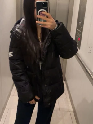Svart pufferjacka från Burberry - Säljer en svart pufferjacka från Burberry med klassiskt rutigt foder i huvan och logga på ärmen. Jackan har lång ärm, dragkedja framtill och en oversized passform som är perfekt för kalla vinterdagar.