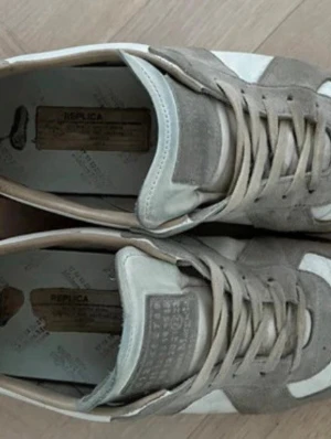 Vita och grå sneakers med mockadetaljer - Snygga sneakers från Maison Margiela i vitt och grått med klassiska mockadetaljer och bruna gummisulor. Skorna har en retroinspirerad design med lågt skaft och snörning framtill. Perfekta för dig som gillar stilrena och tidlösa sneakers. Vid funderingar så är de bara o fråga ✅