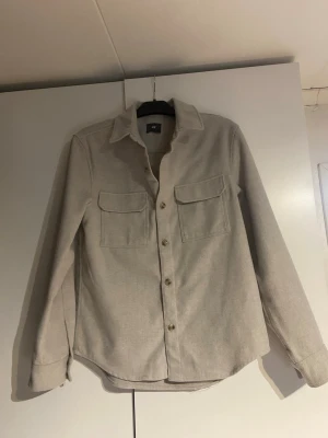 Beige overshirt med fickor - Stilren beige overshirt från H&M med två stora bröstfickor och knäppning framtill. Skjortan har klassisk krage, långa ärmar med manschett och är tillverkad i ett mjukt tyg som ger en avslappnad look.