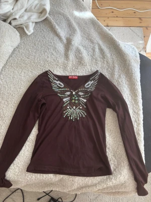 Brun topp med paljettdetaljer Geci Mania - Snygg brun långärmad topp från Geci Mania med v-ringning och stora paljettdetaljer framtill i form av en fjäril. Toppen har en figurnära passform och är perfekt för dig som vill sticka ut med coola detaljer.