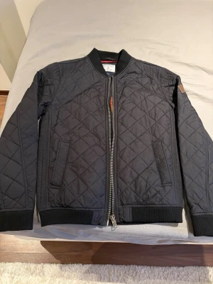 Svart quiltad bomberjacka  - Stilren svart bomberjacka  med quiltat mönster och orange foder. Jackan har ribbade muddar, dragkedja framtill och två snedställda fickor. Perfekt för dig som vill ha en klassisk och cool look.