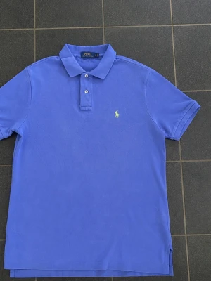 Blå pikétröja från Polo Ralph Lauren - Klassisk blå pikétröja från Polo Ralph Lauren med broderad logga på bröstet. Tröjan har krage, korta ärmar och knäppning med två knappar framtill. Classic fit-modell i mjuk bomull, perfekt för en stilren look.