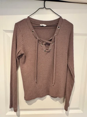Ribbad lila/brun tröja med snörning - Ribbad långärmad tröja i brun nyans från Cubus. Tröjan har snygg snörning framtill med metallöljetter och v-ringad hals. Perfekt för dig som gillar trendiga detaljer och en bekväm passform.