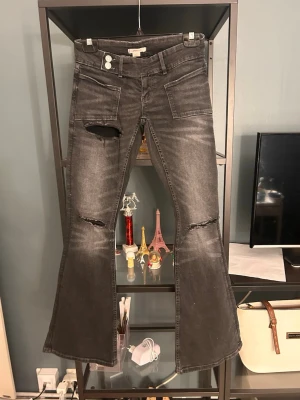 Svarta utsvängda jeans med slitningar - Snygga svarta jeans med utsvängda ben och slitningar på knäna och låret. Jeansen har stora fickor fram och klassiska bakfickor. Perfekta för dig som gillar en edgy och trendig stil. Materialet är denim med en tvättad finish.