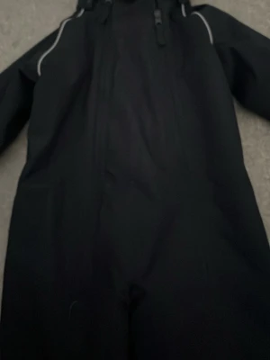Svart overall från Polarn O. Pyret - Svart jumpsuit från Polarn O. Pyret med reflexdetaljer och dragkedja framtill. Gjord i slitstarkt, återvunnet material, vattentät och vindtät med tejpade sömmar. Andas bra och har mjukt foder. Perfekt för utomhusaktiviteter för barnen eller en skidtur