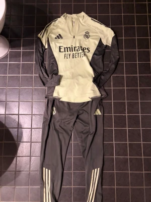 Real Madrid träningsset Adidas - Snyggt Real Madrid träningsset från Adidas med ljusgul tröja och mörkgrå ärmar samt byxor. Tröjan har halv dragkedja, klubbmärke och Emirates Fly Better-tryck. Byxorna är mörkgrå med ljusgula detaljer och tre ränder längs benen. Perfekt för fotbollsträning.