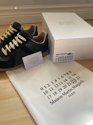 Maison Margiela svarta sneakers - Maison Margiela sneakers i svart läder och mocka med beige detaljer och vita snören. Klassisk låg modell med gummisula och diskret logotyp på sulan. Kommer med originalkartong och dustbag. Perfekta för dig som gillar stilrena och exklusiva sneakers. Skick 7,5/10 har en liten slitning på insidan av ena.