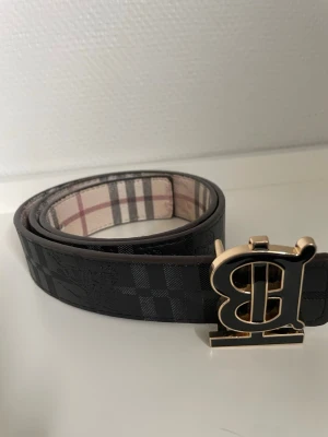  bälte med guldspänne - Snyggt bälte med klassiskt rutigt mönster i beige, svart och rött. Bältet har ett stort guldigt B-spänne och är tillverkat i skinn. Perfekt accessoar för att lyfta din outfit.
