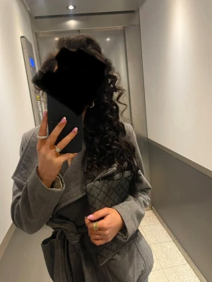 Grå omlottkappa med knytskärp - Snygg grå kappa i omlottmodell med knytskärp i midjan. Kappan har en klassisk krage och raka ärmar, perfekt för en stilren look. Materialet ser ut att vara mjukt och ger en elegant känsla. Passar perfekt till höst och vinter.