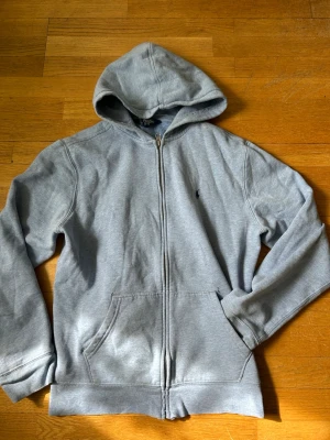 Ljusblå hoodie från Polo Ralph Lauren - Klassisk grå hoodie med dragkedja från Polo Ralph Lauren. Tröjan har huva, två fickor framtill och den ikoniska broderade loggan på bröstet. Perfekt för en chill och avslappnad stil. Tillverkad i mjukt bomullsmaterial. Passar 175-179 S.