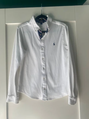 Vit skjorta från Polo Ralph Lauren - Klassisk vit skjorta från Polo Ralph Lauren med lång ärm och knappar framtill. Skjortan har en broderad blå logga på bröstet och button-down krage. Perfekt för en stilren look och tillverkad i mjuk bomull. För barn, ålder 14-16 år. 