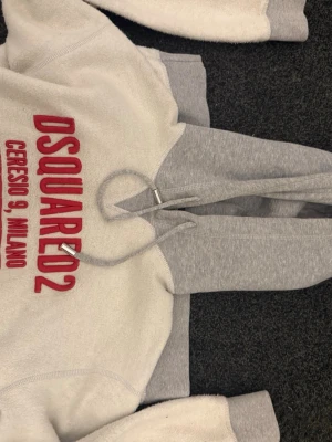 DSQUARED2 vit och grå hoodie - Vit och grå hoodie från DSQUARED2 med röd broderad text 'DSQUARED2 Ceresio 9, Milano' på bröstet. Hoodien har huva och en stor magficka. Tillverkad i mjukt material, köpt på nk i Göteborg och är limeted edition så går inte att få tag på någonstans, nypris ca 5/6 tusen men säljer den för 3 tusen, fick den i julklapp men den var för stor så därför jag säljer 🫶🏼🫶🏼