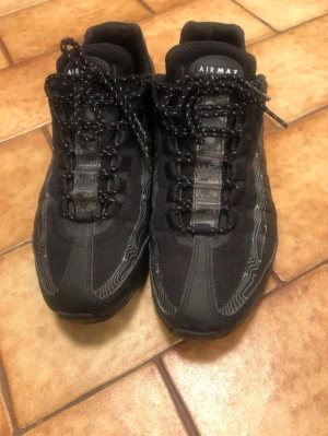 Svarta Nike Air Max sneakers - Snygga svarta Nike Air Max sneakers med reflekterande detaljer och mönstrade svarta skosnören. Ovandelen är i mesh och syntetmaterial för en sportig look. Klassisk Air Max-logga på plösen och coola linjer längs sidorna.