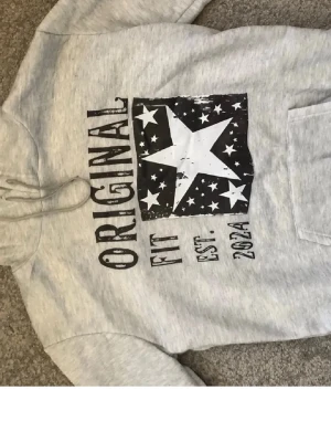 Grå hoodie med stjärntryck - Snygg grå hoodie från Original Fit med stor vit stjärna och svart/vitt tryck på bröstet. Huva med dragsko och magficka framtill. Perfekt för en avslappnad och cool stil. Trycket har texten 'ORIGINAL FIT EST. 2024' och flera små stjärnor.