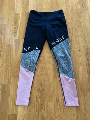 Färgglada leggings från H&M Sport - Snygga leggings från H&M Sport med svart överdel, gråmelerad mitt och ljusrosa ben. De har tryckt text på låren och är perfekta för träning eller chill. Materialet är mjukt och stretchigt syntet för maximal komfort.
