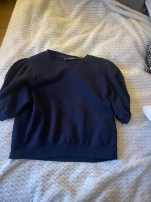 Mörkblå topp från Gina Tricot - Mörkblå topp från Gina Tricot med korta puffärmar och rund halsringning. Tjockare ribbad kant vid ärmslut och nederkant ger en snygg passform. Perfekt att styla med jeans eller kjol för en trendig look.