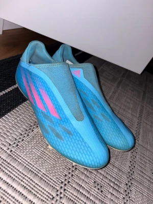 Blåa Adidas fotbollsskor utan snörning - Säljer ett par blåa Adidas fotbollsskor med rosa ränder på sidan. Skorna är utan snörning och har en strumpliknande passform. Yttermaterialet är syntetiskt och sulan är anpassad för spel på gräs. Perfekta för dig som vill ha stilrena och snabba skor på planen.