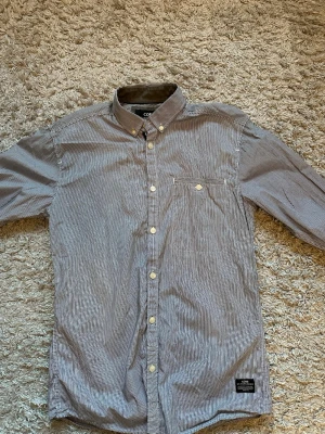 Randig skjorta  - Snygg skjorta med smala vita och svarta/mörkblå ränder. Klassisk krage med knappar, bröstficka och vita knappar framtill. Perfekt för en stilren look och passar till många olika outfits.
