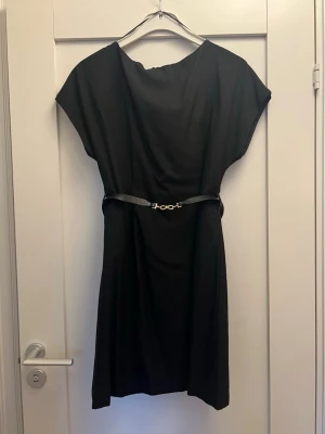 Svart kortklänning med bälte - En stilren svart kortklänning med lös passform och ett smalt bälte i midjan. Klänningen har korta ärmar och rund halsringning, perfekt för dig som gillar enkel och klassisk stil. Materialet känns mjukt och följsamt.