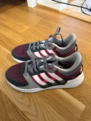 Adidas sneakers vinröd och grå - Snygga Adidas sneakers i vinrött och grått med klassiska vita ränder på sidan. Ovandelen är i mesh och mockaliknande material, med grå snörning och vit sula. Skön passform och sportig look som passar perfekt till streetwear. Helt ny och oanvänd! Storlek 36 men kommer i stor storlek!
