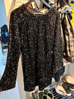 Svart paljettklänning med långa ärmar - Glittrig svart kortklänning täckt av paljetter, med rund halsringning och långa ärmar. Perfekt för dig som vill sticka ut och glänsa på festen. Klänningen har en figurnära passform och är tillverkad i ett syntetmaterial. Köpt till nyår förra året men använde inte den💕