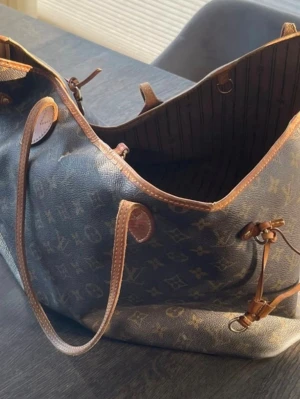 Louis Vuitton Neverfull Monogram väska - Säljer en klassisk Louis Vuitton Neverfull shoppingväska i brunt monogrammönstrat canvasmaterial med läderdetaljer. Väskan har långa axelband, öppet huvudfack och randigt innerfoder. Perfekt för dig som gillar lyxiga och rymliga väskor.