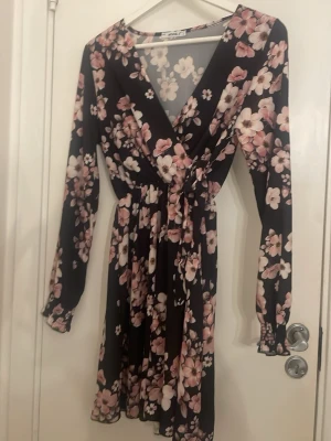 Svart blommig omlottklänning - Snygg svart omlottklänning med rosa och vita blommor. Klänningen har långa ärmar med resår vid ärmslut och markerad midja. V-ringad framtill och lätt utsvängd kjol. Perfekt för dig som gillar romantiska mönster och sköna material.